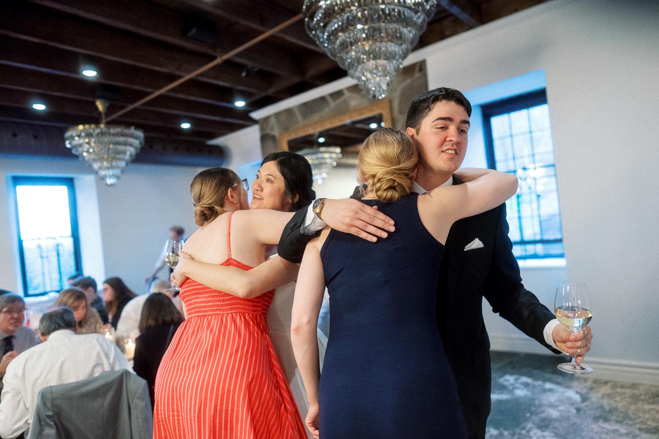 Guests hug warmly during an indoor engagement celebration with chandeliers and seated diners.