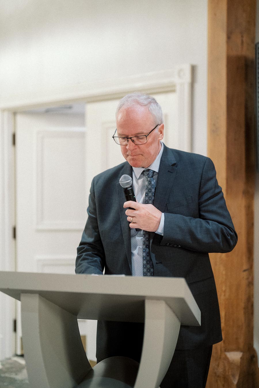 Man in a dark suit speaking into a microphone at a lectern indoors