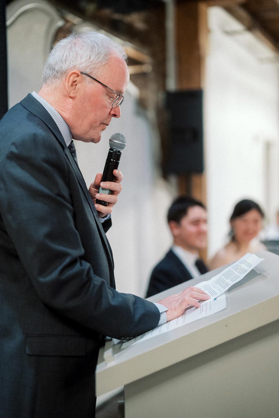 Man in a suit speaking into a microphone at a podium with a couple behind him