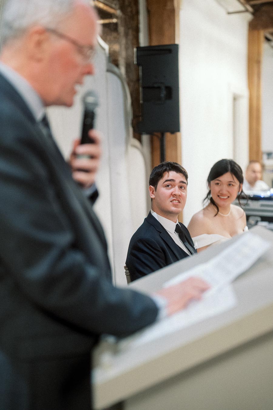 Couple in wedding attire listening to a speaker at a reception table