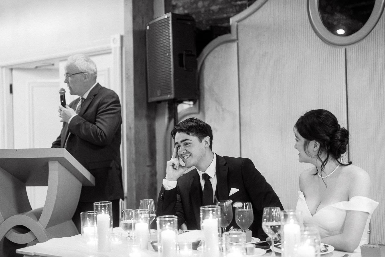 Couple at a candlelit reception table listening as a man gives a speech