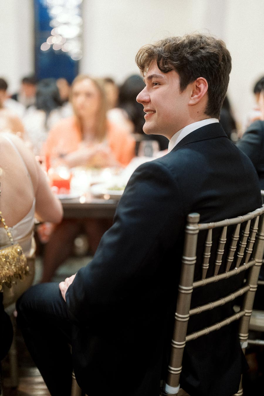 Man in a black suit smiling while seated at a formal dinner table