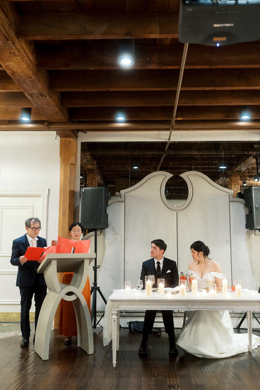 Couple seated at a reception table while two speakers read at a podium