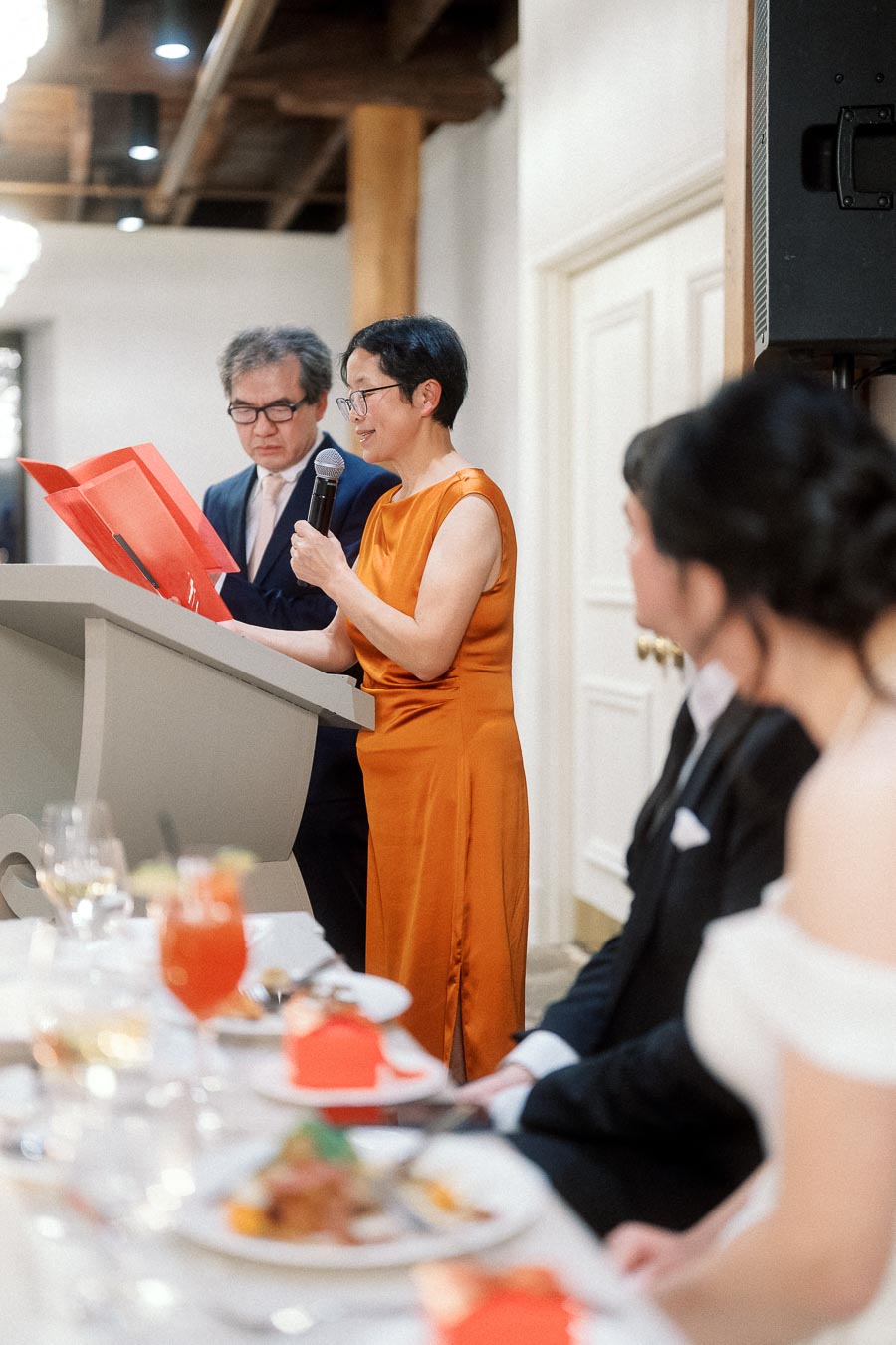 Woman in orange dress gives a speech at a formal reception podium.