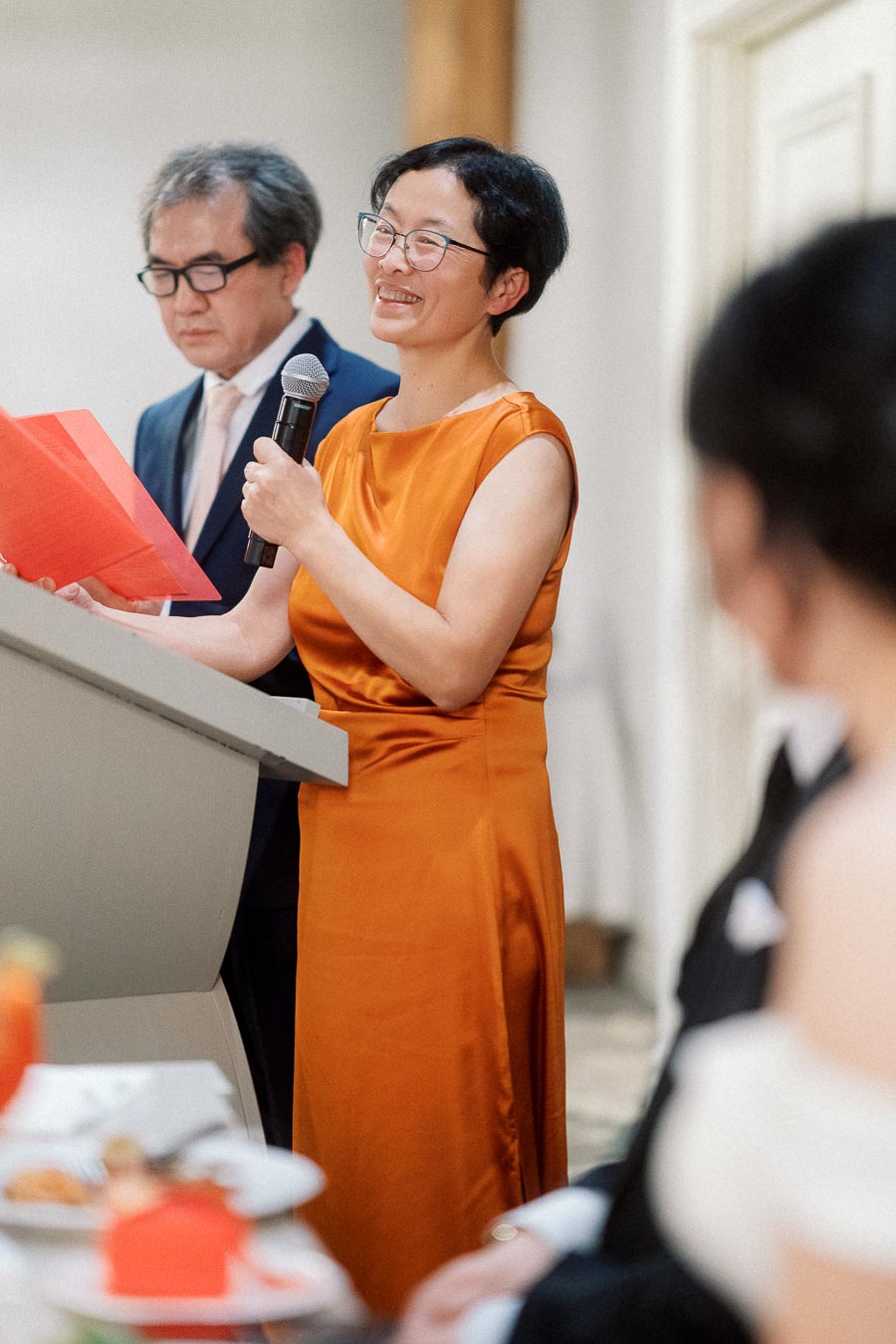 Woman in an orange dress gives a speech at a formal indoor event.
