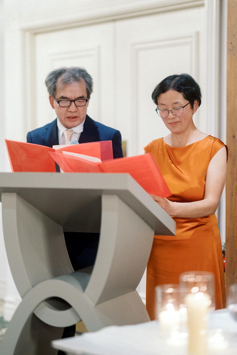 Two adults reading from red folders at a lectern during an indoor ceremony