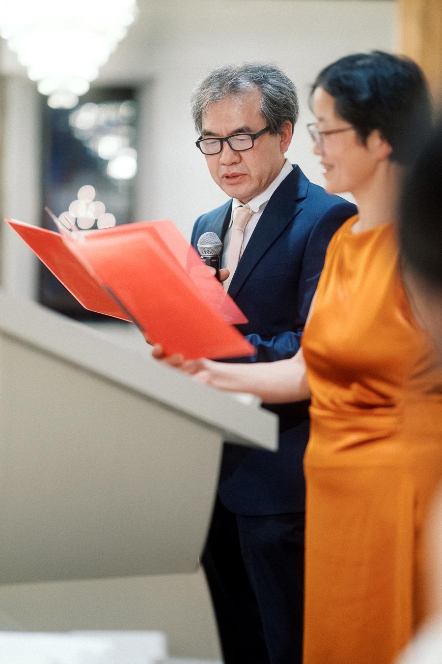 Man in a suit reads from a red folder while holding a microphone indoors