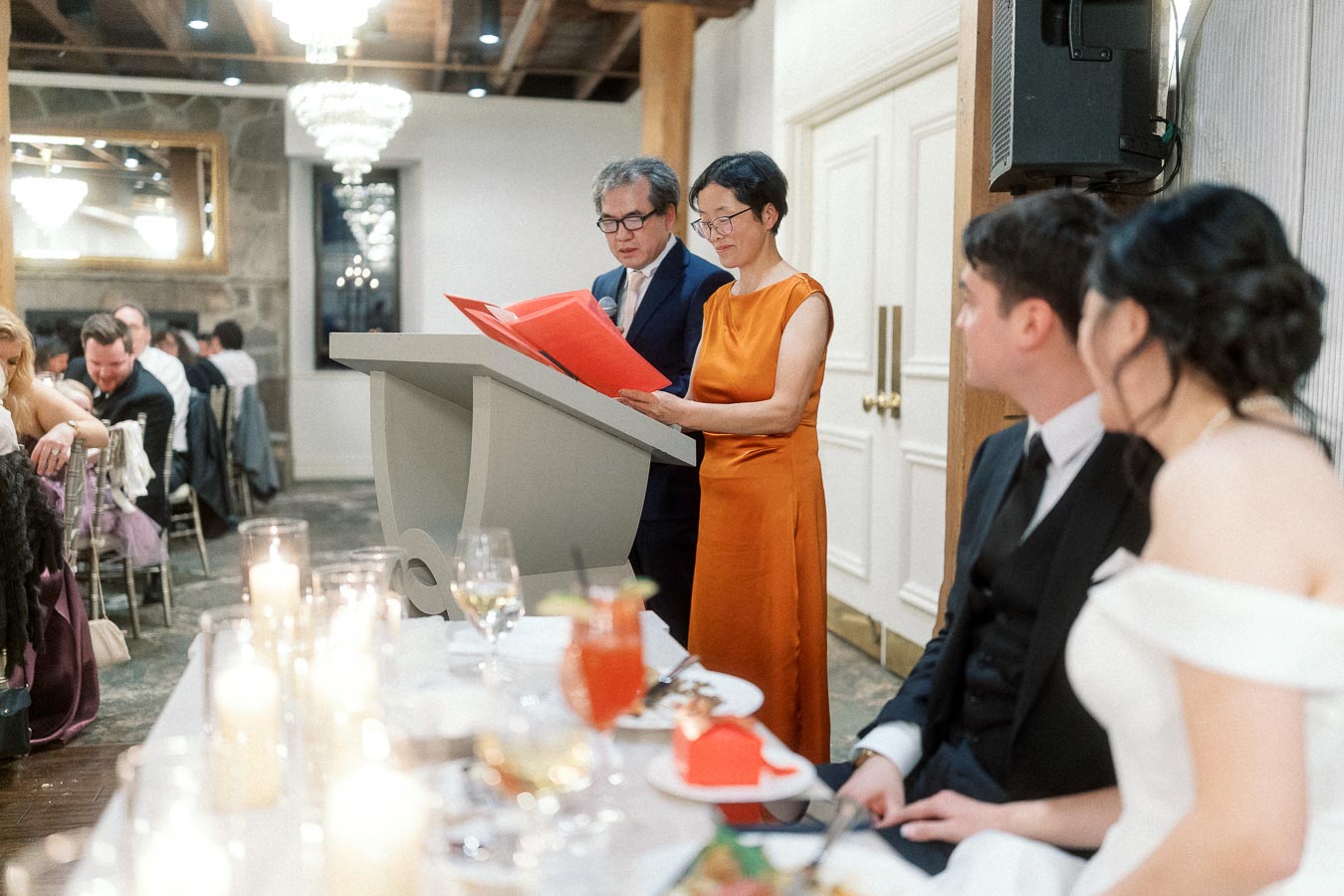 Two guests read from a red folder at a reception podium as a seated couple listens.