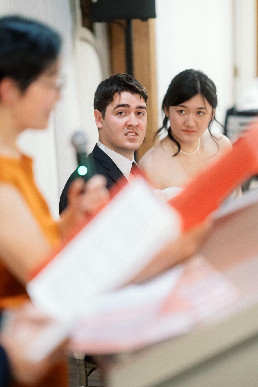 Formally dressed couple seated indoors while someone reads from papers in the foreground.