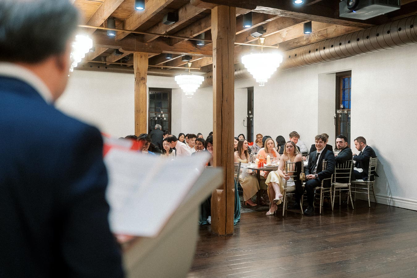 Speaker at a formal indoor event addressing seated guests at round tables