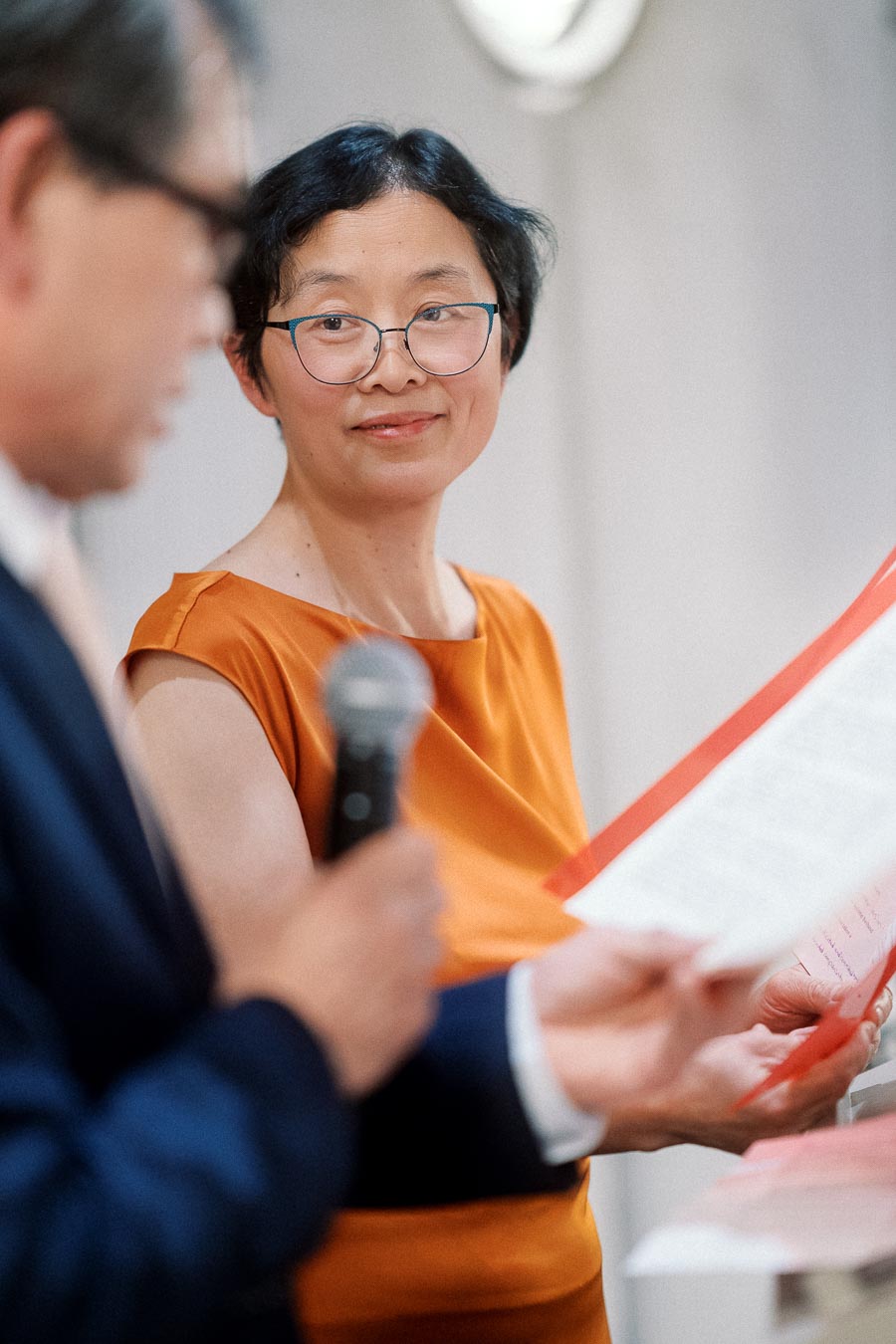 Woman in an orange dress listens as a man reads from papers into a microphone.