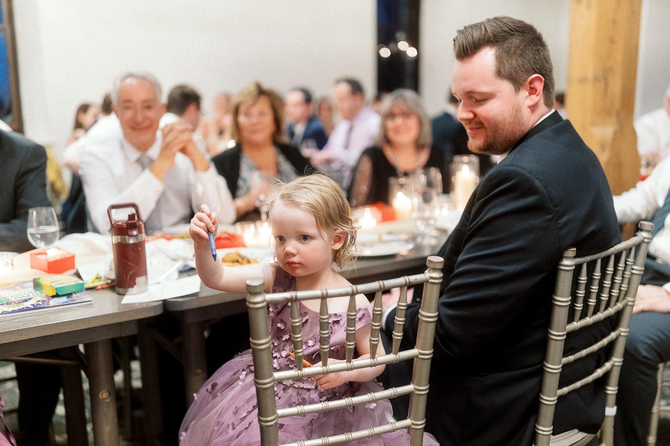 Young child in a purple dress sits beside a man at a candlelit reception table.