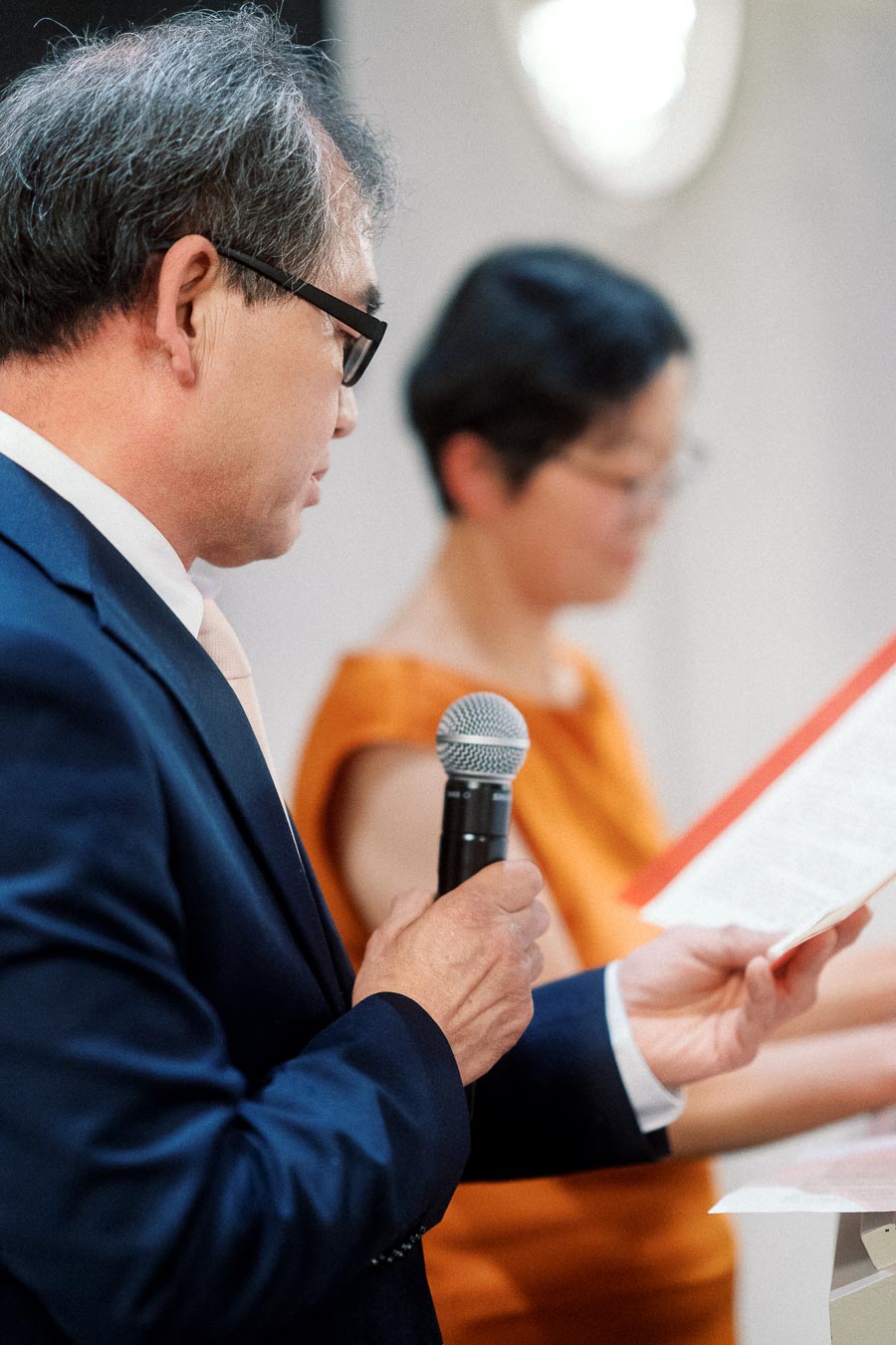 Man in a navy suit reads from paper while holding a microphone.