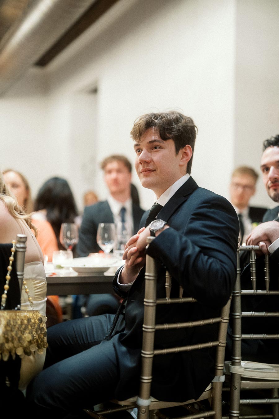 Man in a dark suit seated among guests at a formal indoor event