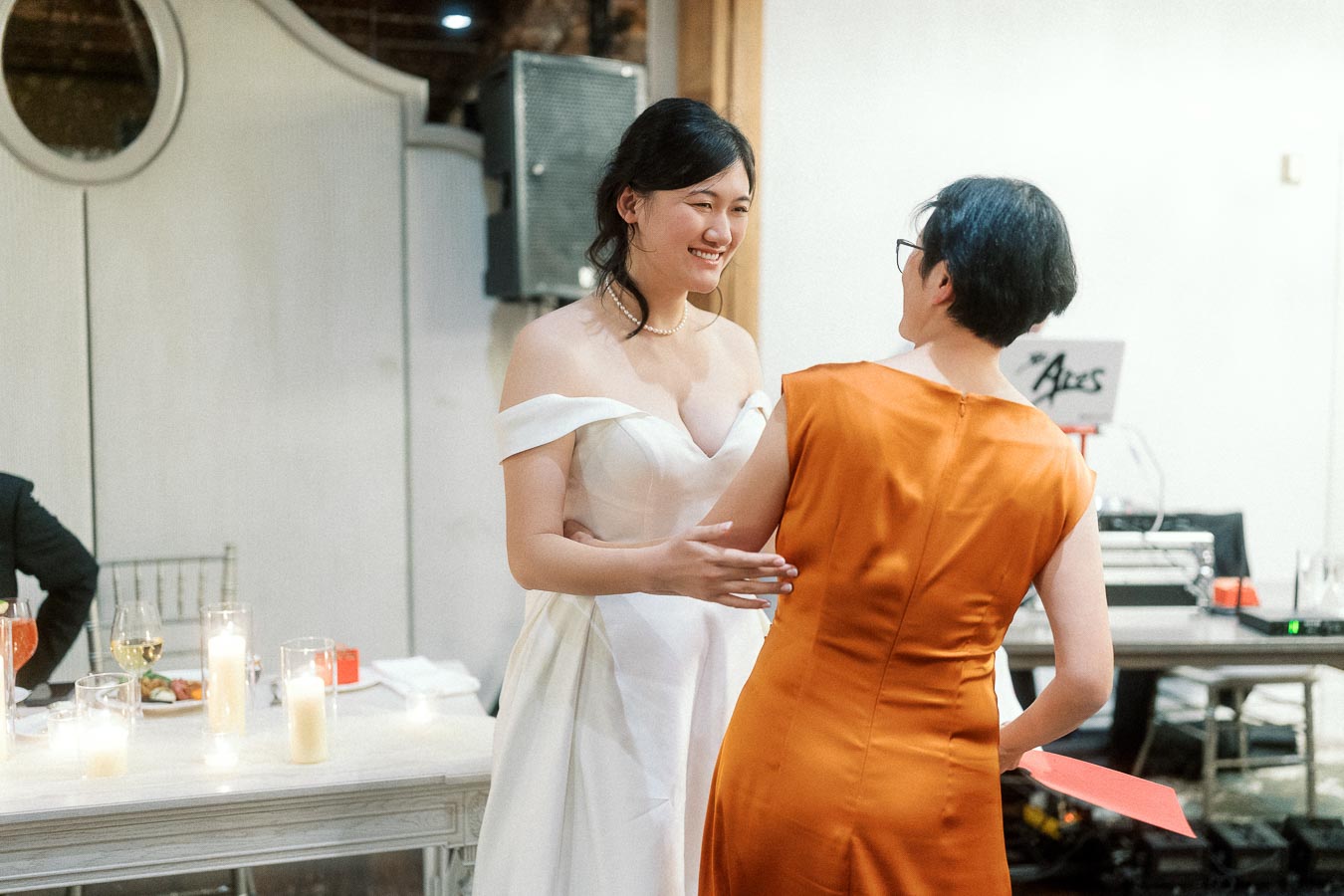 Bride in white gown smiling while greeting a guest in an orange dress indoors