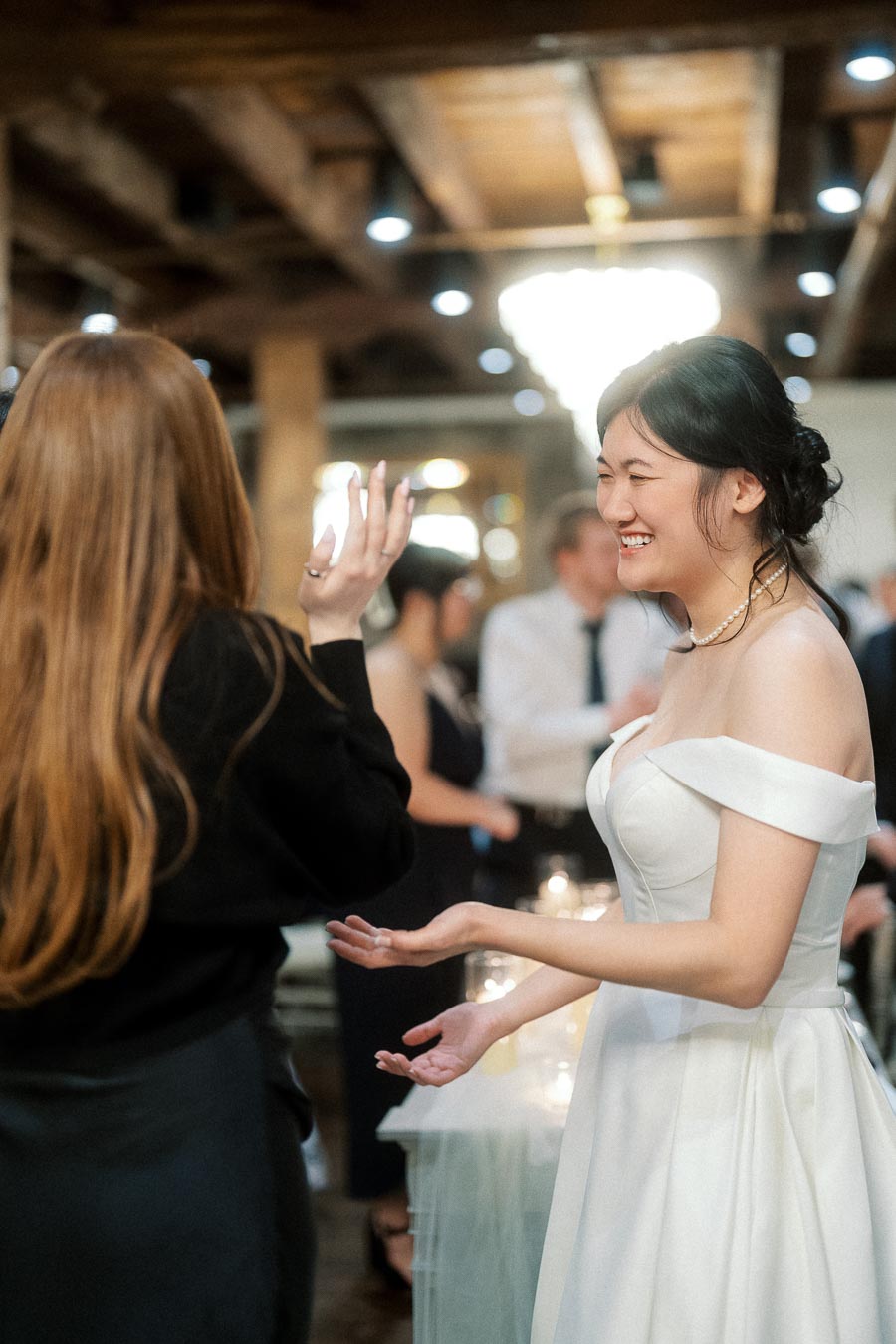 Woman in a white dress laughs while talking with another guest at an indoor celebration.
