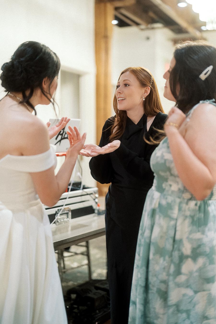 Three women talking animatedly indoors, including one in a white off-shoulder dress.