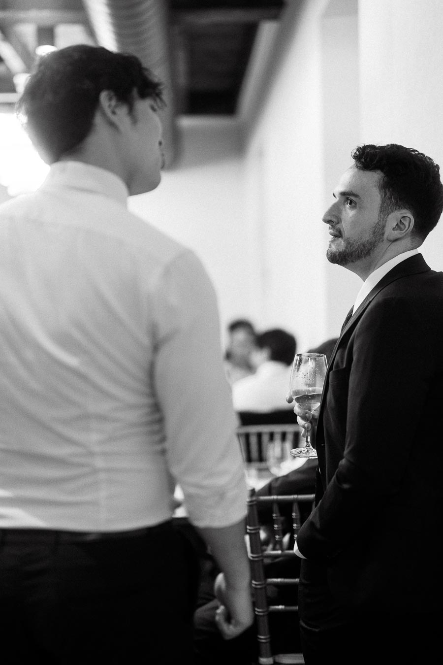 Two men talking indoors, one in a suit holding a wine glass.