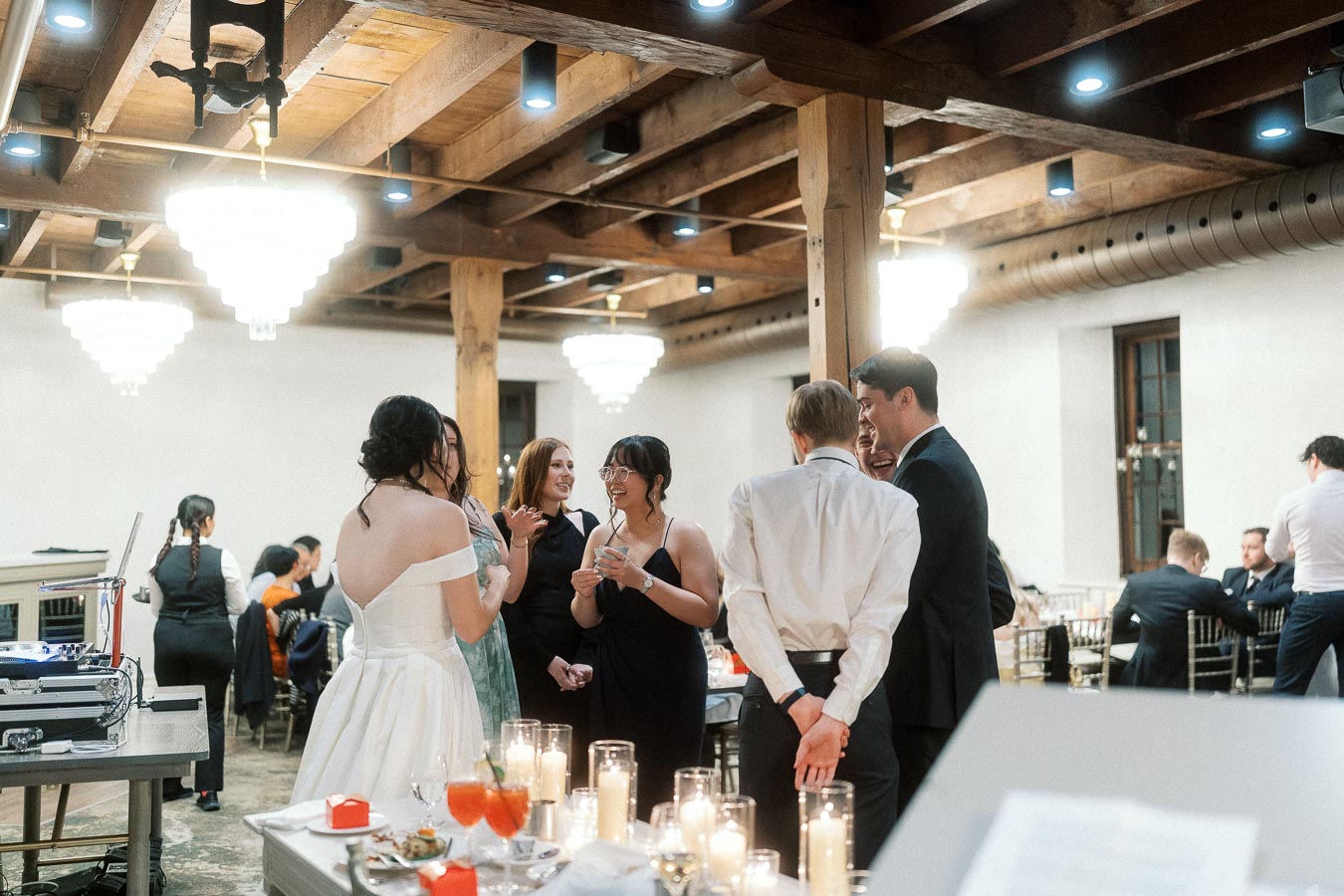 Couple and guests chatting indoors beside candlelit tables under wooden beams.