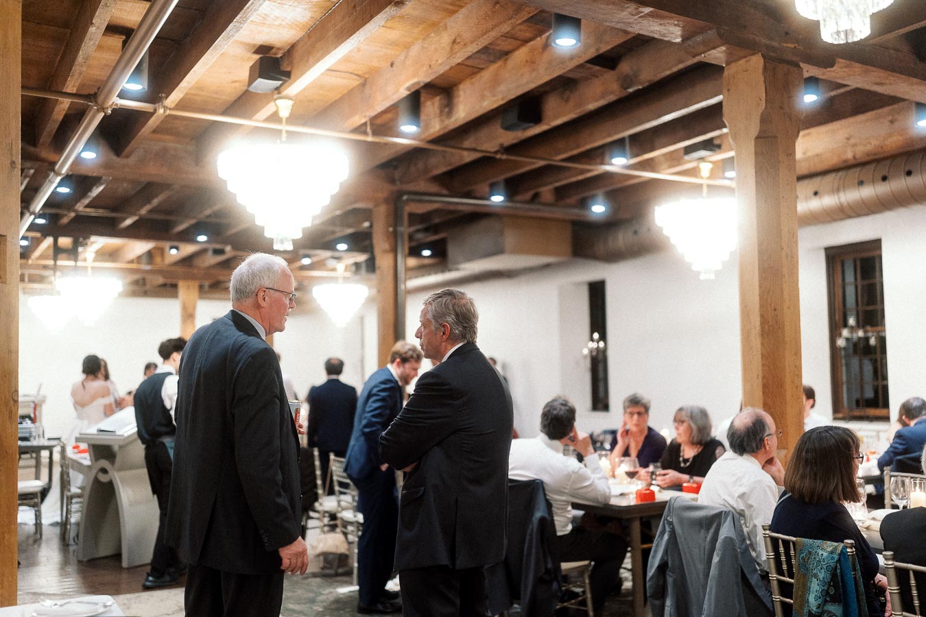 Guests in formal attire mingle and dine in a warm wood-beamed event space.