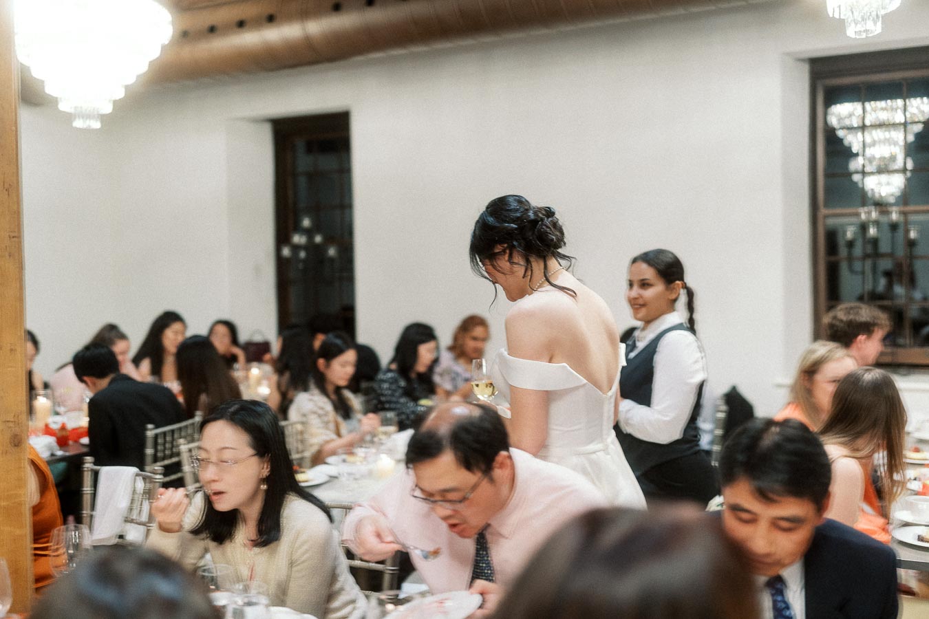 Bride in an off-the-shoulder dress mingles with guests during an indoor reception dinner.