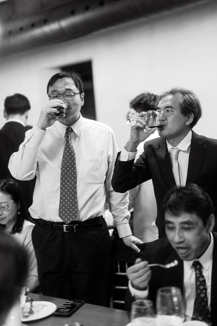 Two men in dress shirts and suits drink from glasses at a reception table.