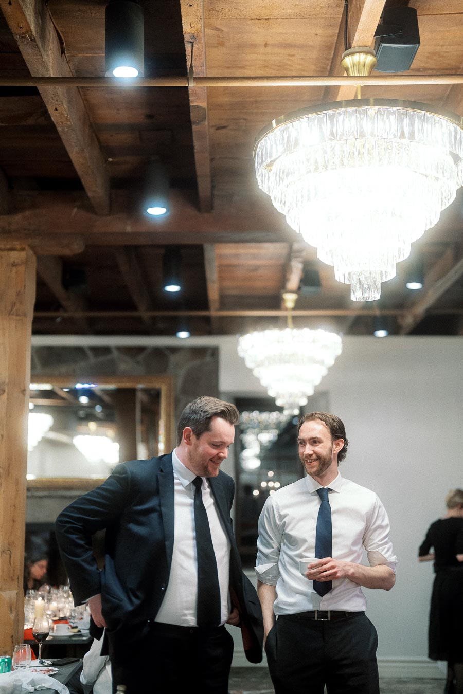 Two men in dress shirts and ties smiling beneath chandeliers indoors