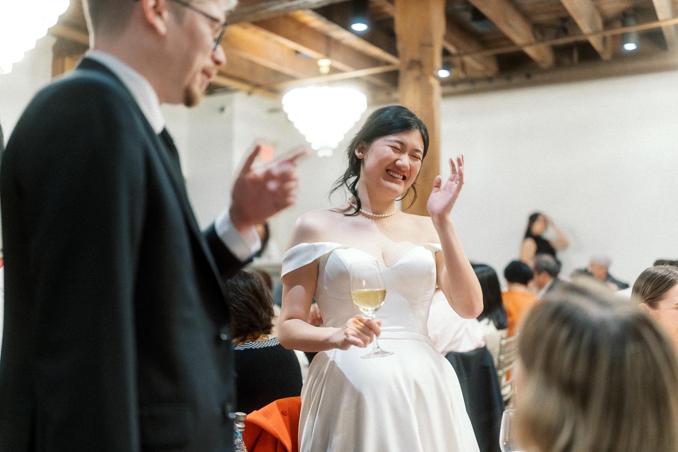 Woman in a white off-shoulder dress laughs while holding a wine glass indoors.