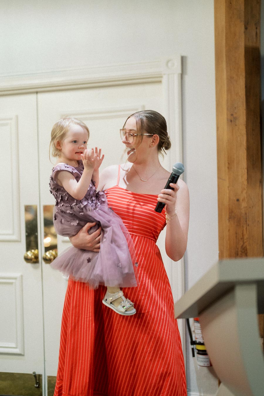 Woman in a red striped dress holds a toddler while carrying a microphone indoors.