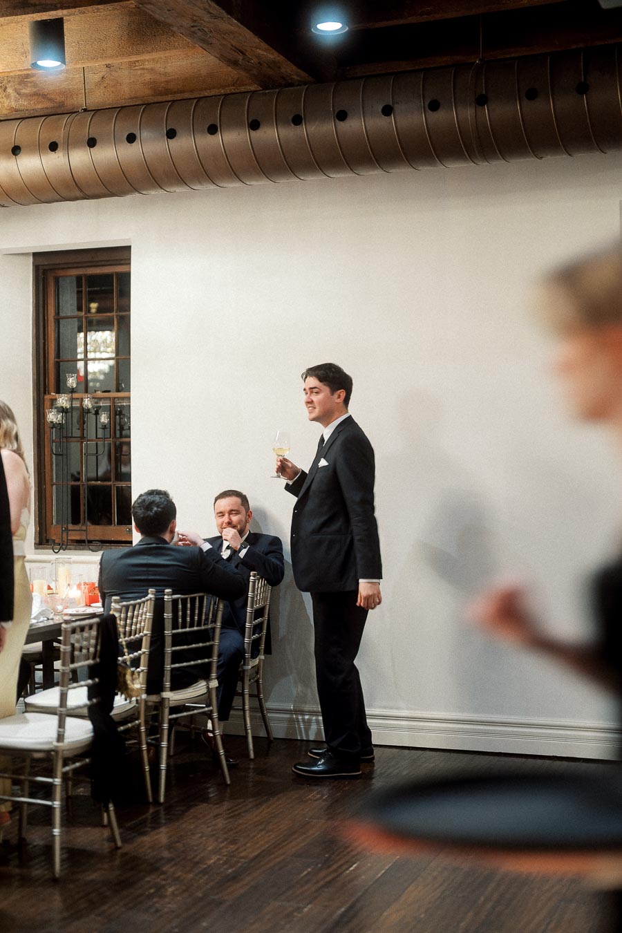 Man in a black suit holding a drink beside seated guests at an indoor reception