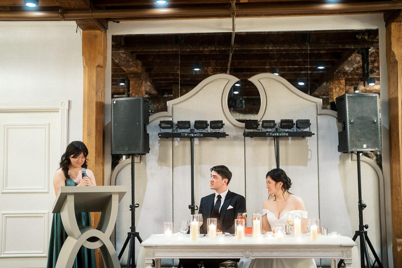 Woman gives a speech while a formally dressed couple sits at a candlelit head table.