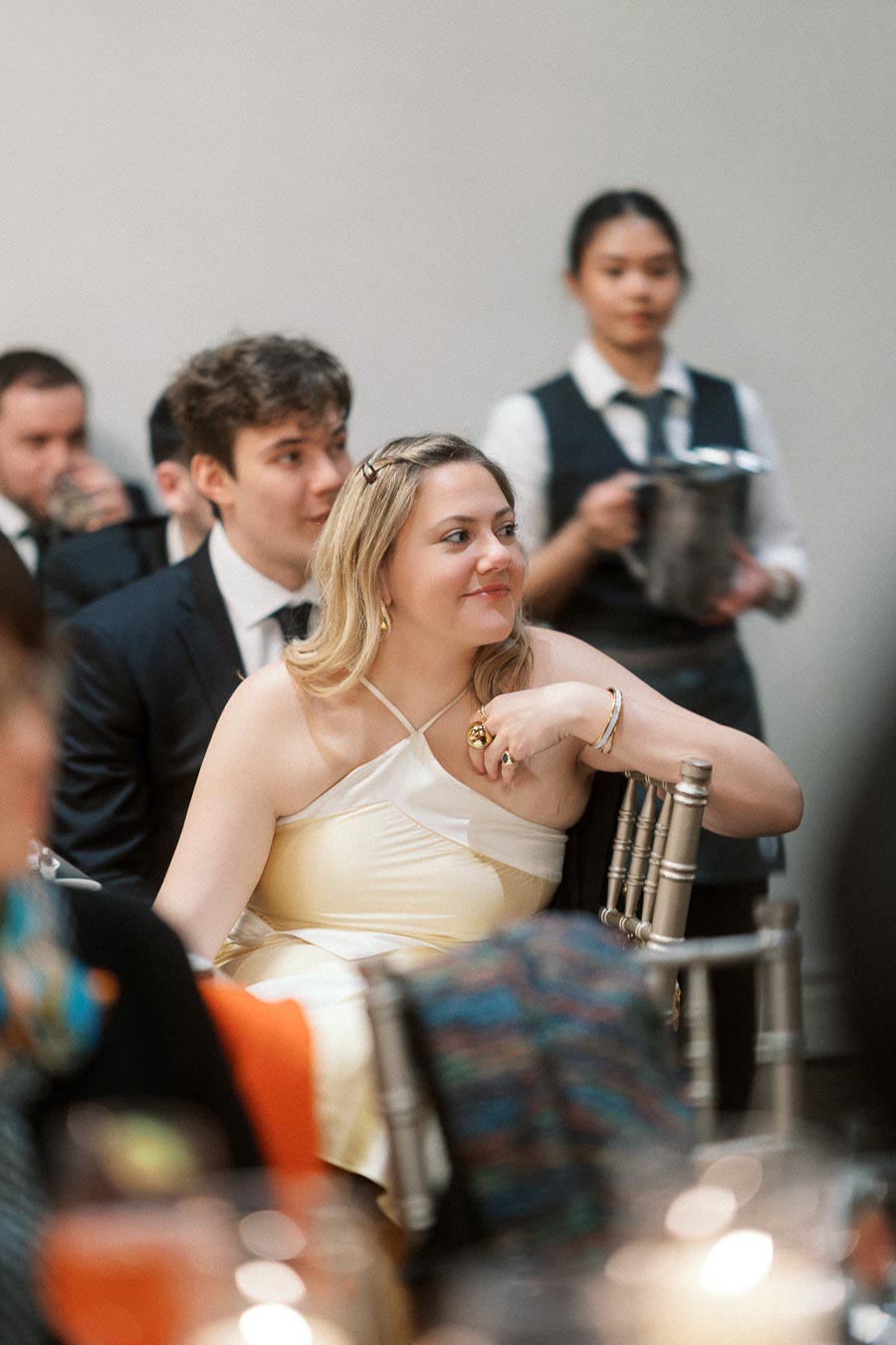 Woman in a cream dress seated at an indoor event with guests and a server nearby