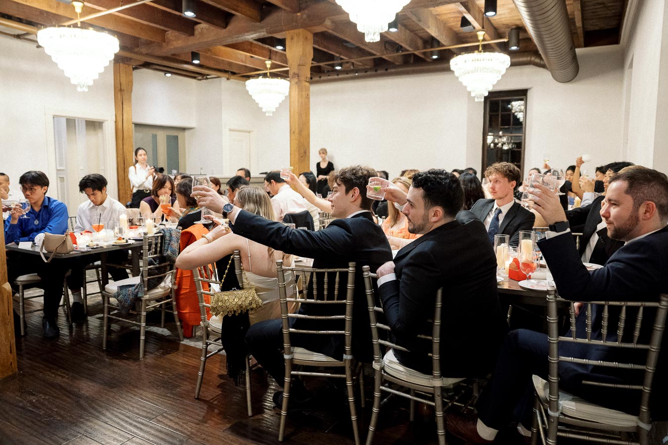 Guests seated at banquet tables raise glasses for a toast in a chandelier-lit reception room.