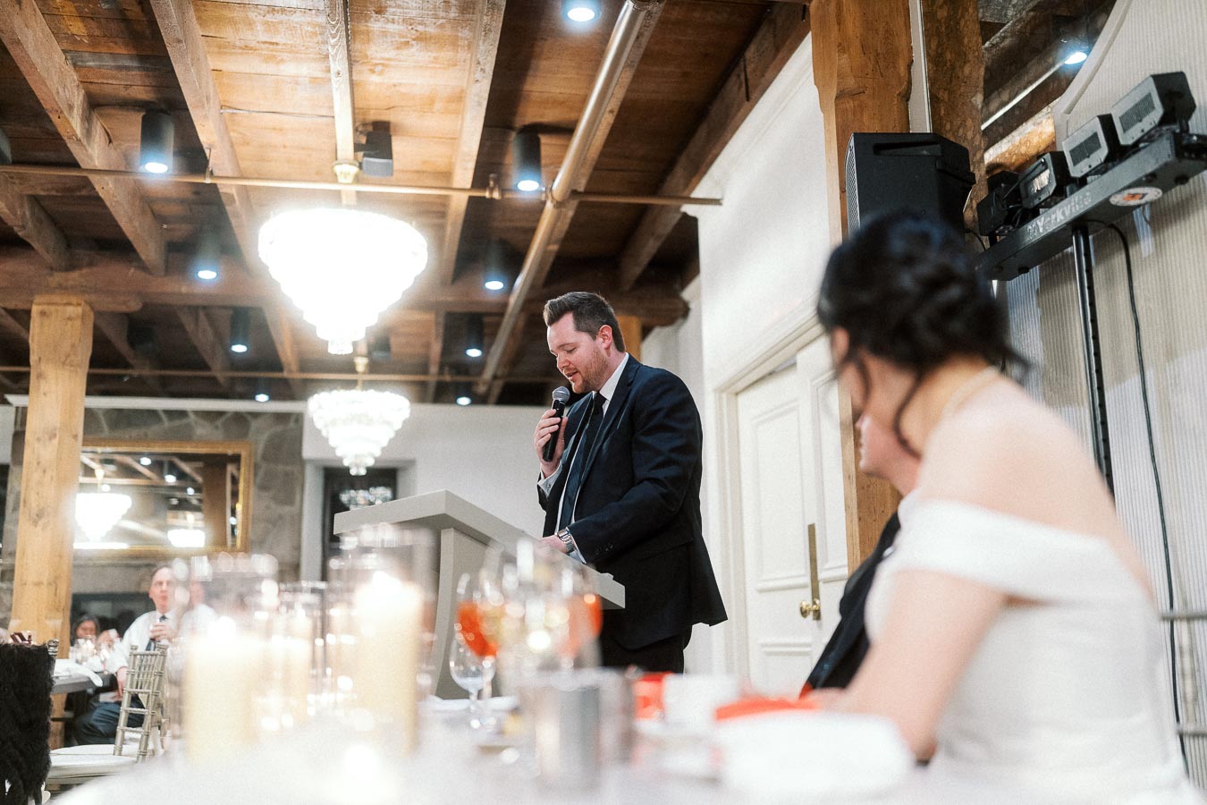 Man in a suit gives a microphone speech at an indoor reception.
