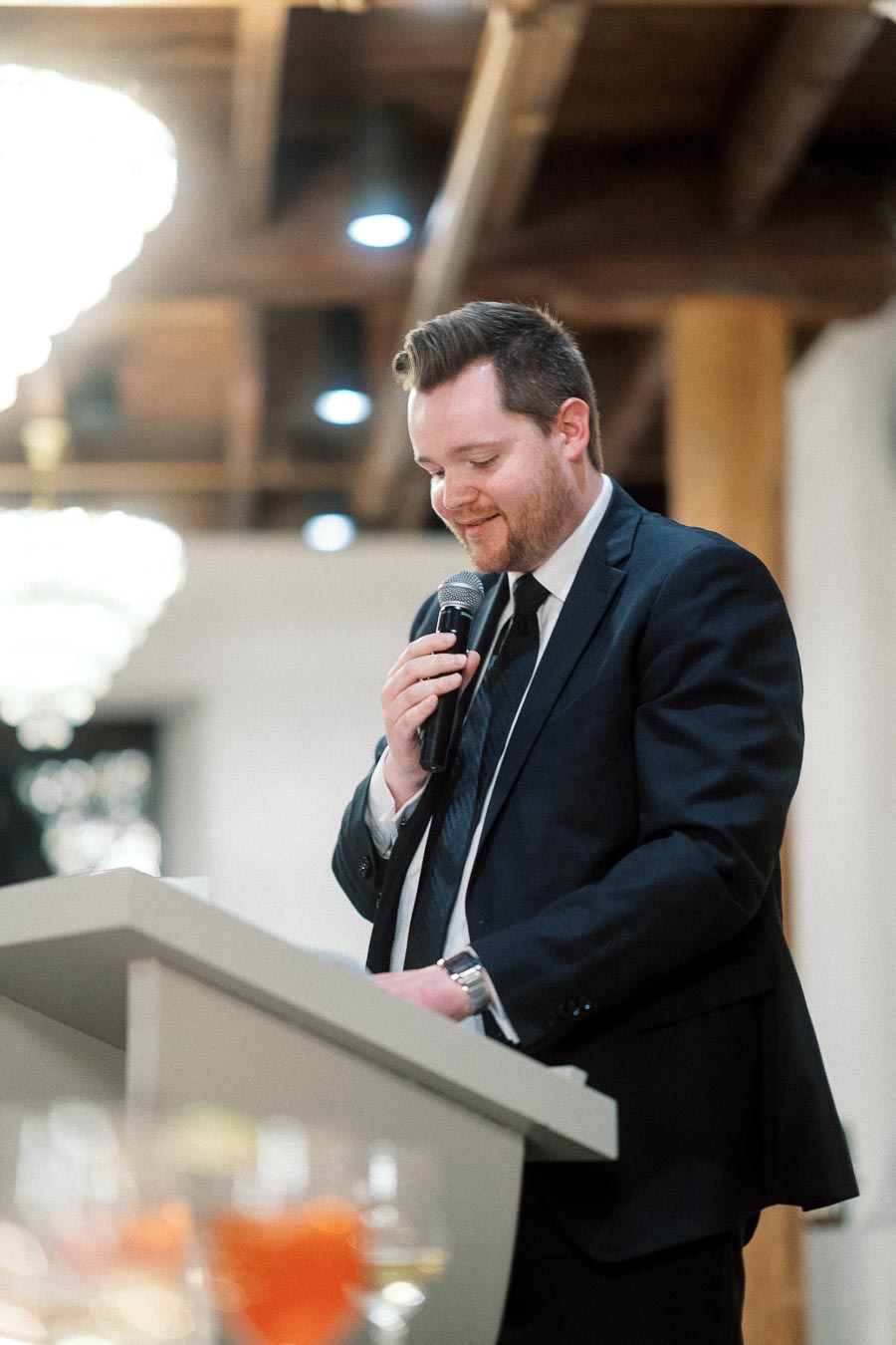 Man in a black suit speaking into a microphone at a podium indoors