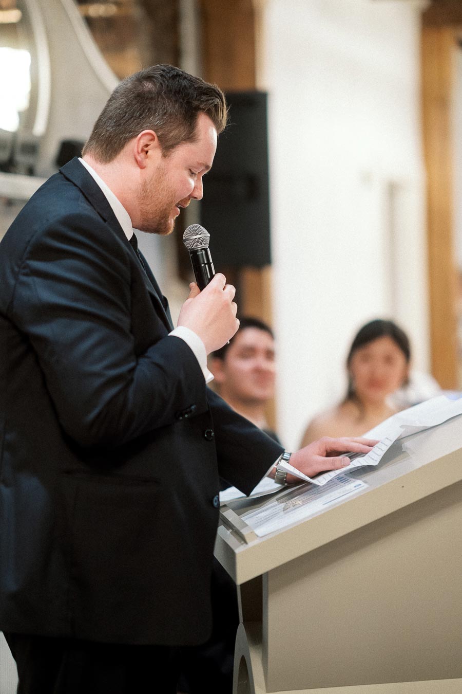 Man in a black suit reads from notes while speaking into a microphone.