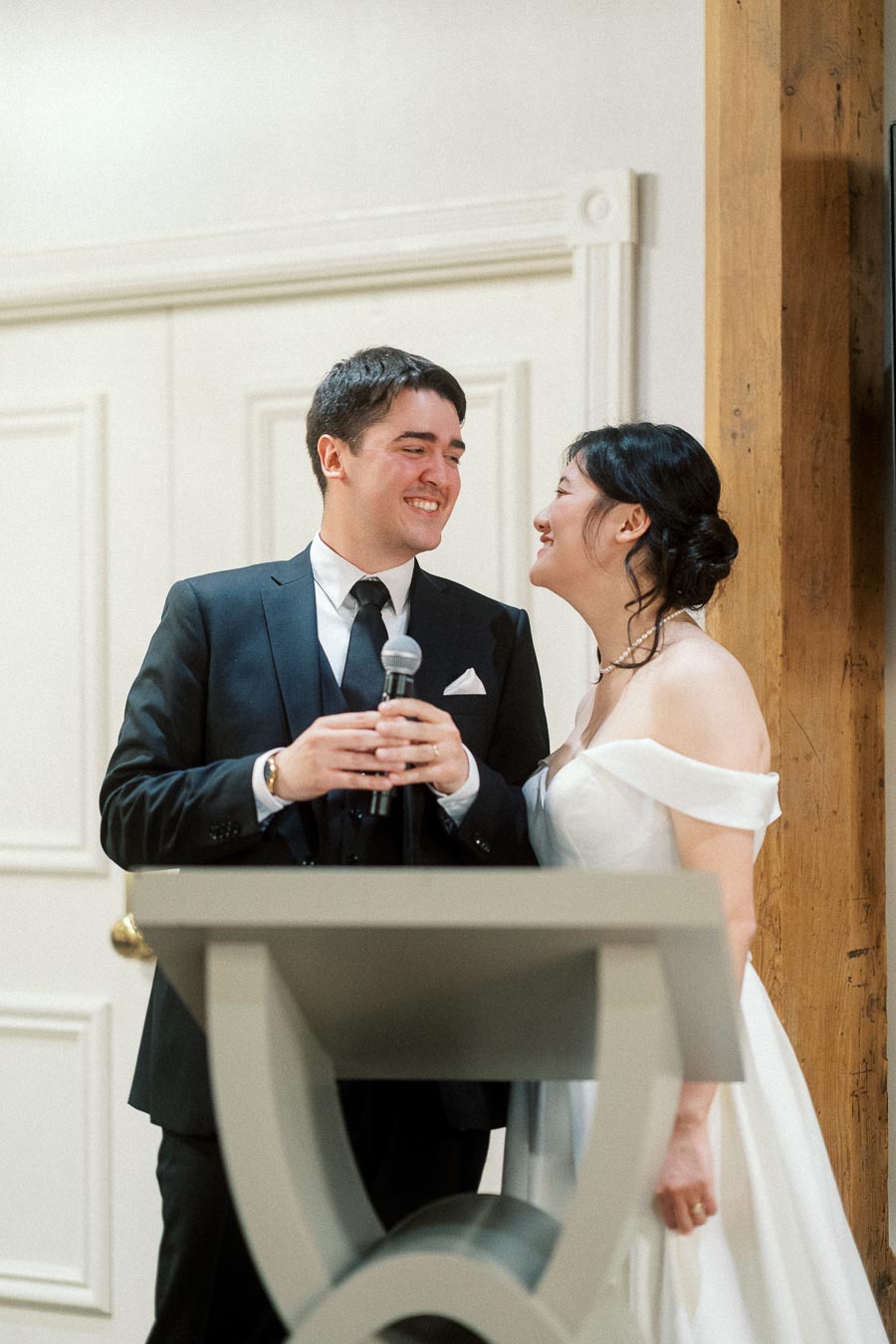 Couple in formal wedding attire smiling together at a podium with a microphone