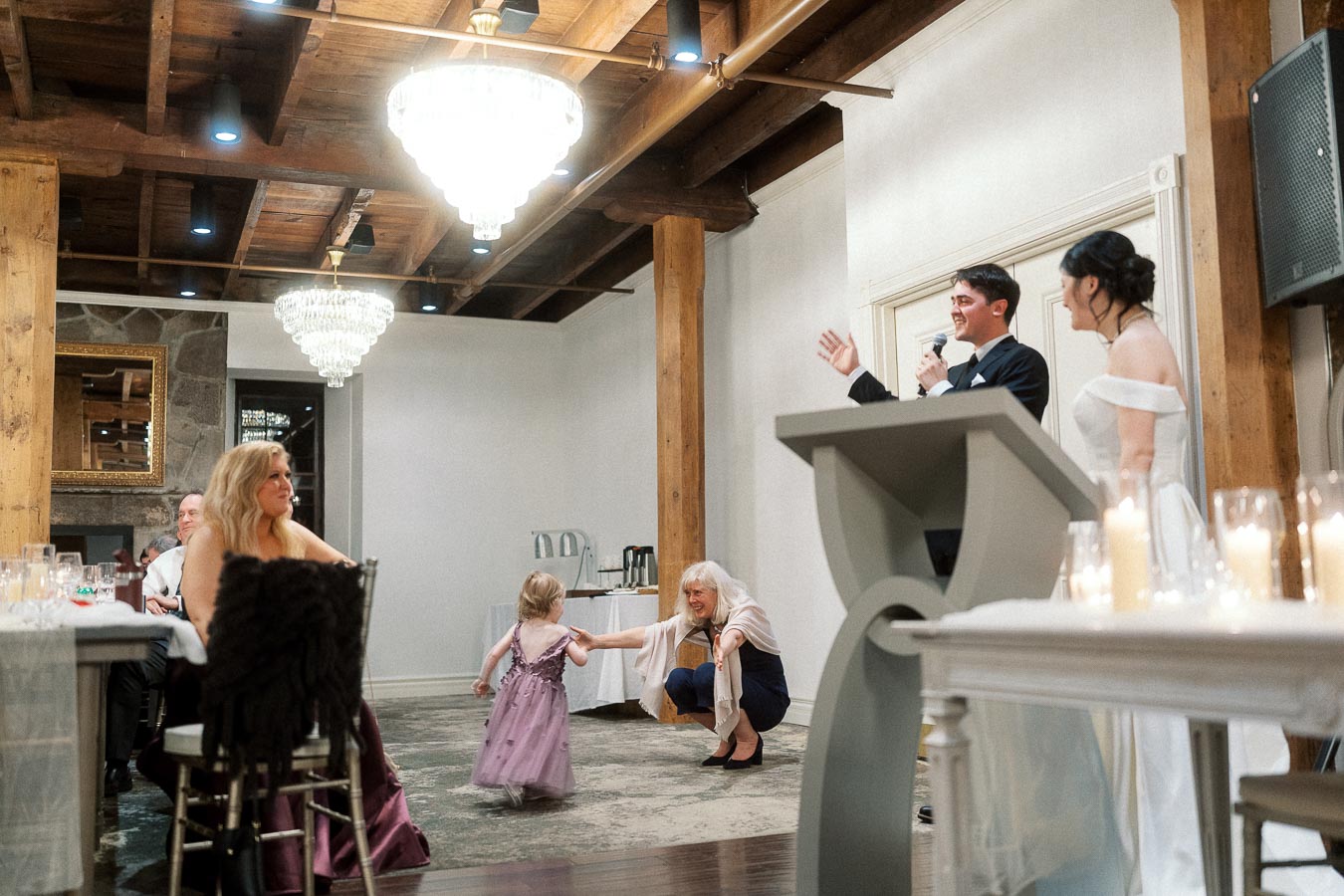 Groom gives a speech beside the bride as a child walks across the reception room.