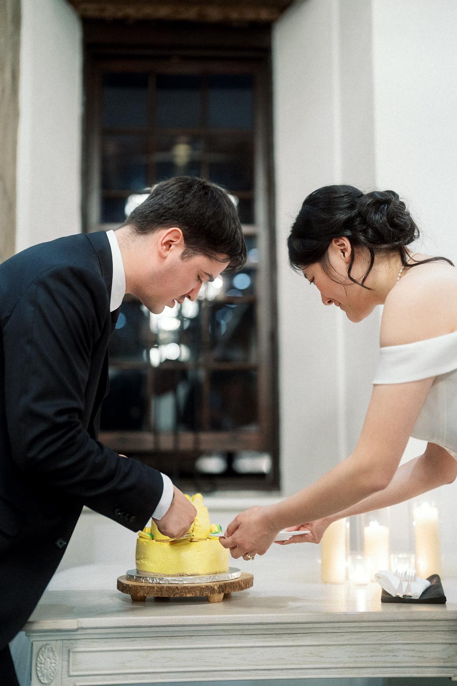 Couple in formal outfits cutting a yellow cake together at a candlelit table.