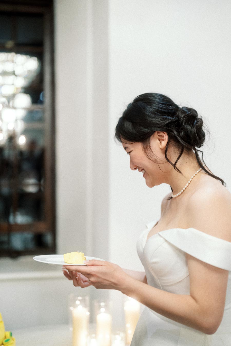 Woman in an off-shoulder white dress smiling while holding a small plate of cake