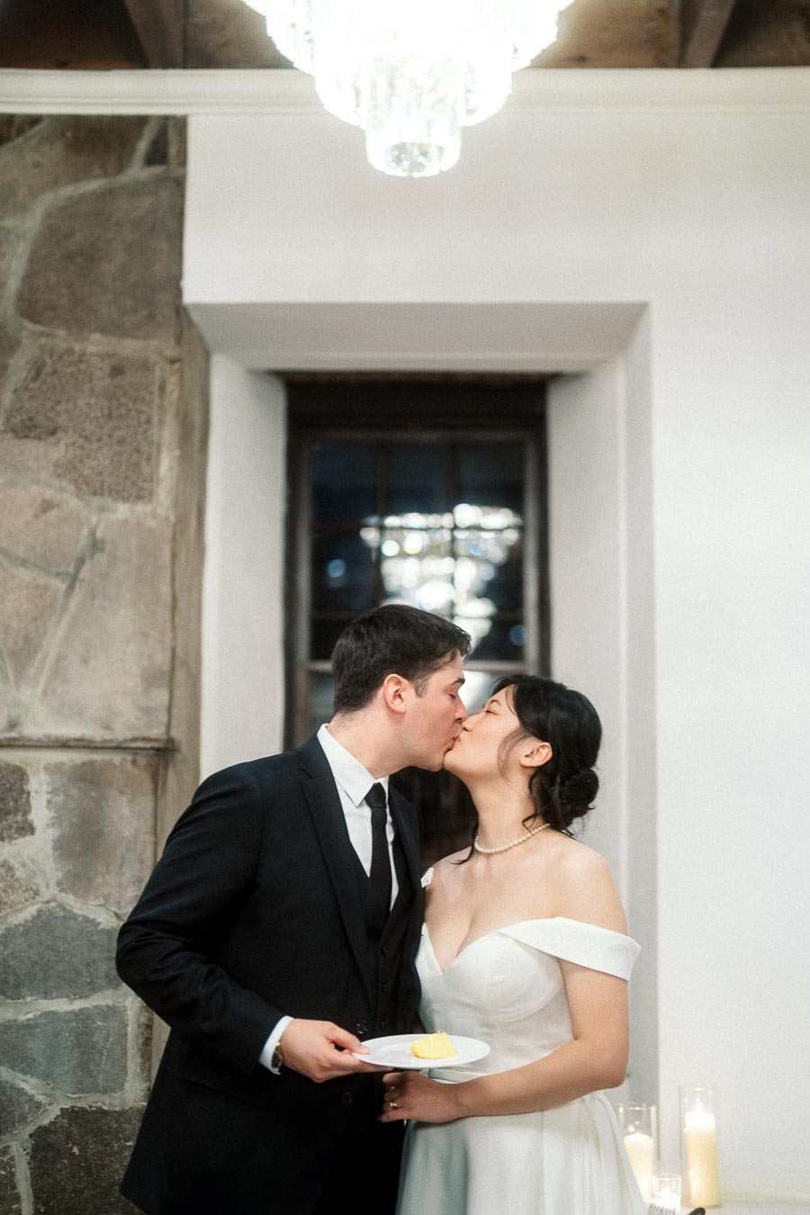 Couple in formal attire kissing indoors while holding a small dessert plate.