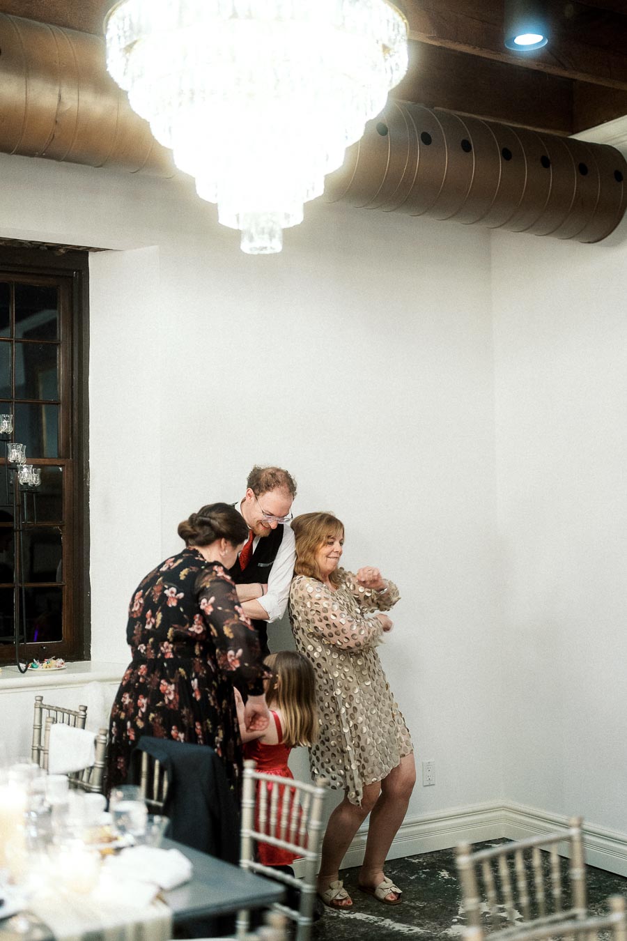 Guests dance together indoors beneath a large chandelier at a celebration.