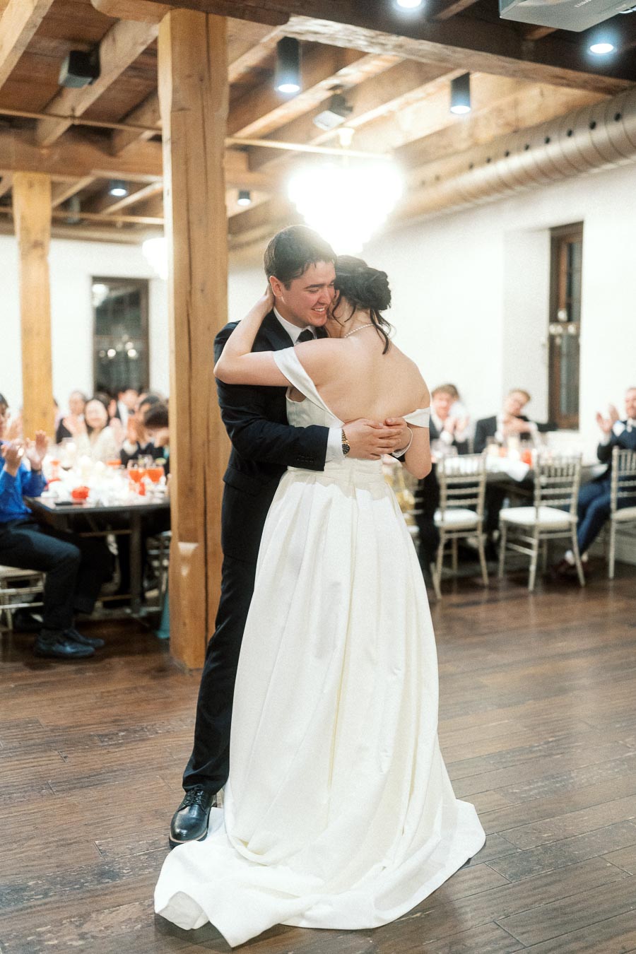 Couple embracing during a dance at an indoor reception with seated guests applauding