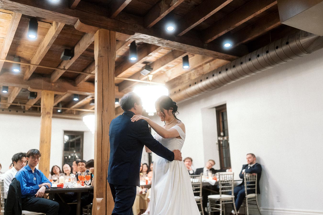 Couple dancing together at an indoor reception with seated guests watching.