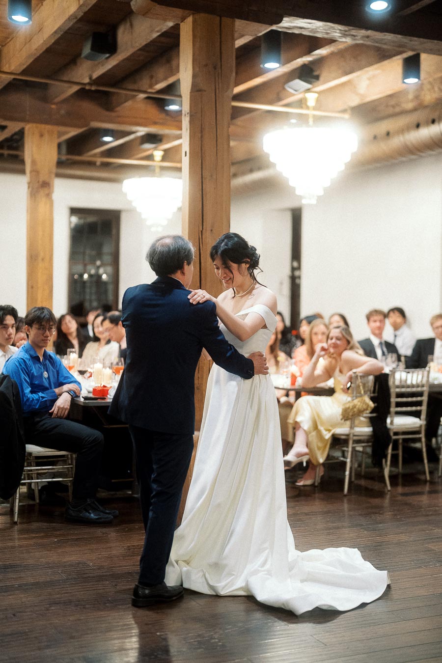 Bride in a white gown dances with an older man during an indoor reception.