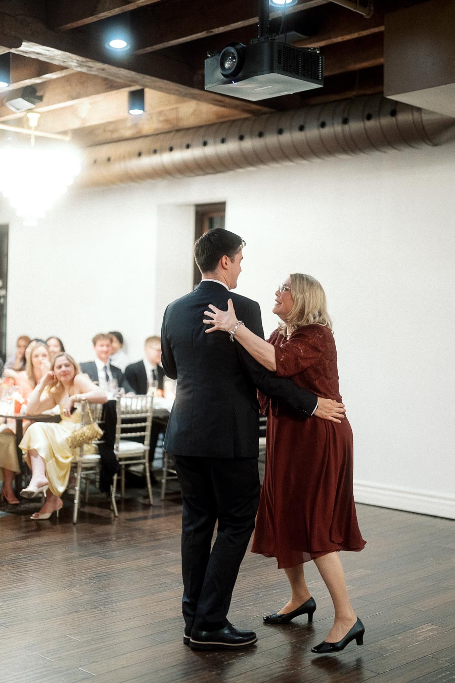 Man in a suit dances with a woman in a burgundy dress at an indoor reception.