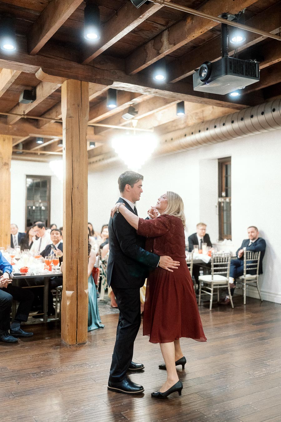 Two people slow dancing on a wooden floor during an indoor celebration.
