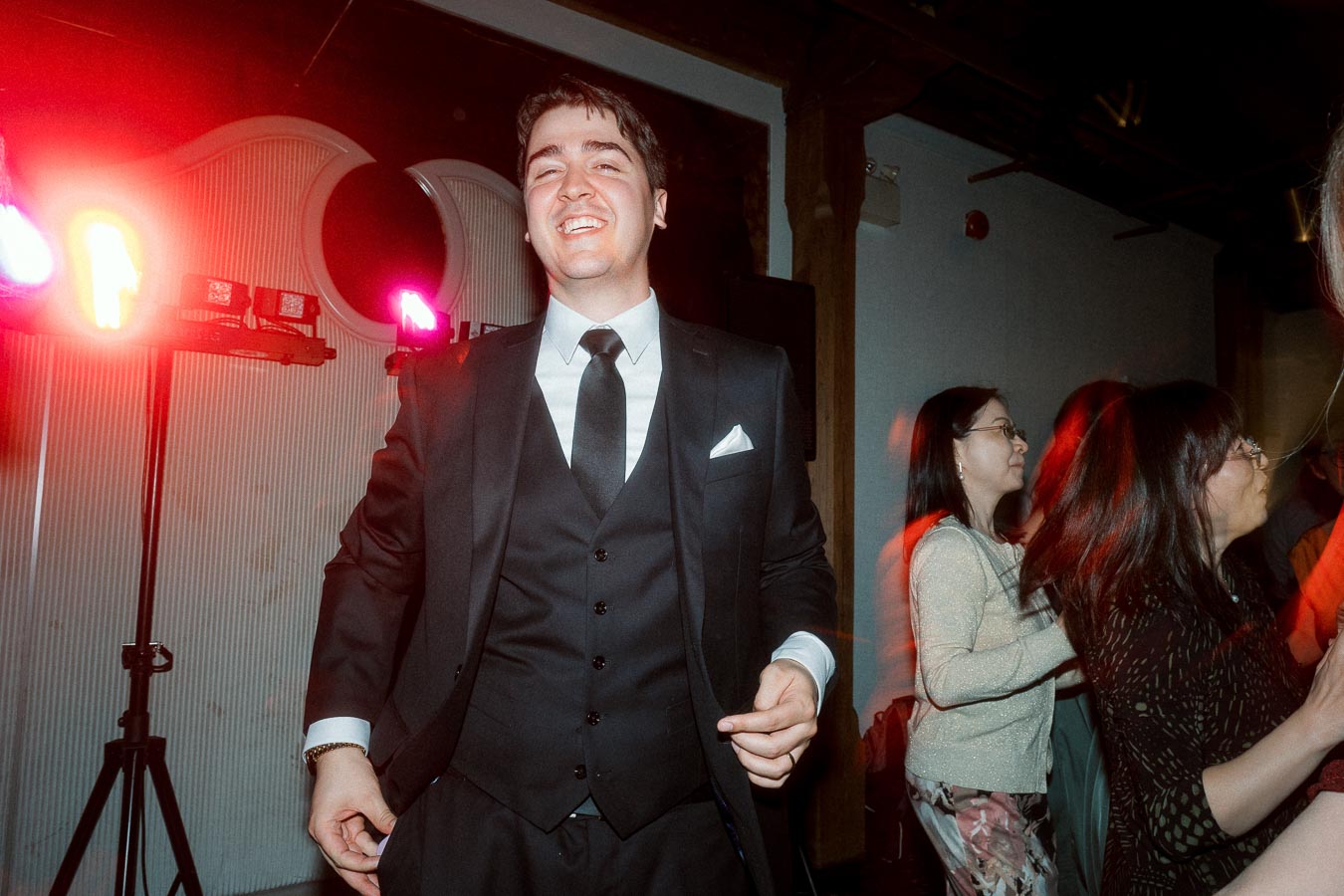 Groom in a black suit smiling on a dance floor with red party lights