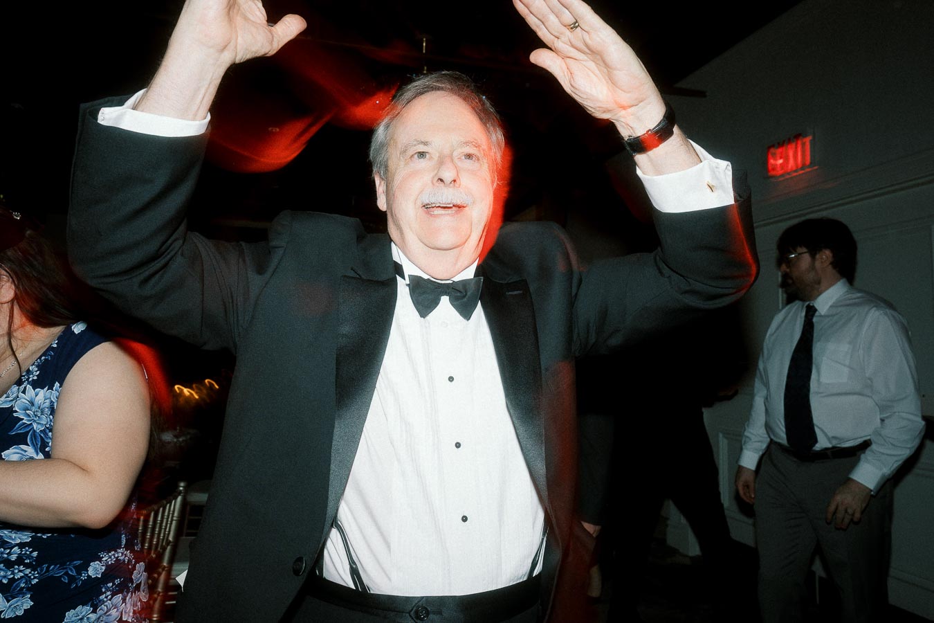 Man in a tuxedo dancing with raised hands at an indoor celebration