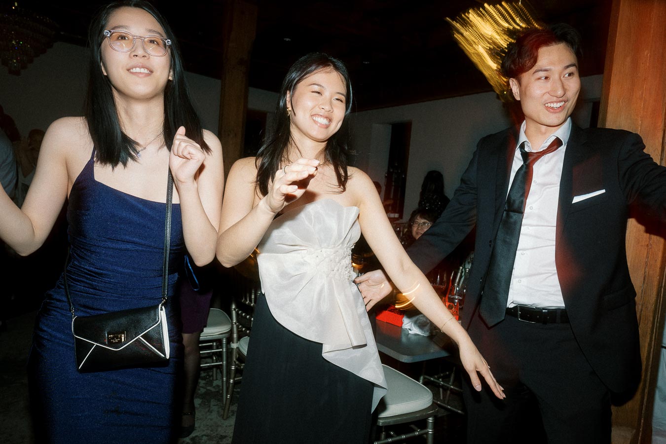 Three guests dancing indoors at a formal celebration with chairs and warm lights behind them